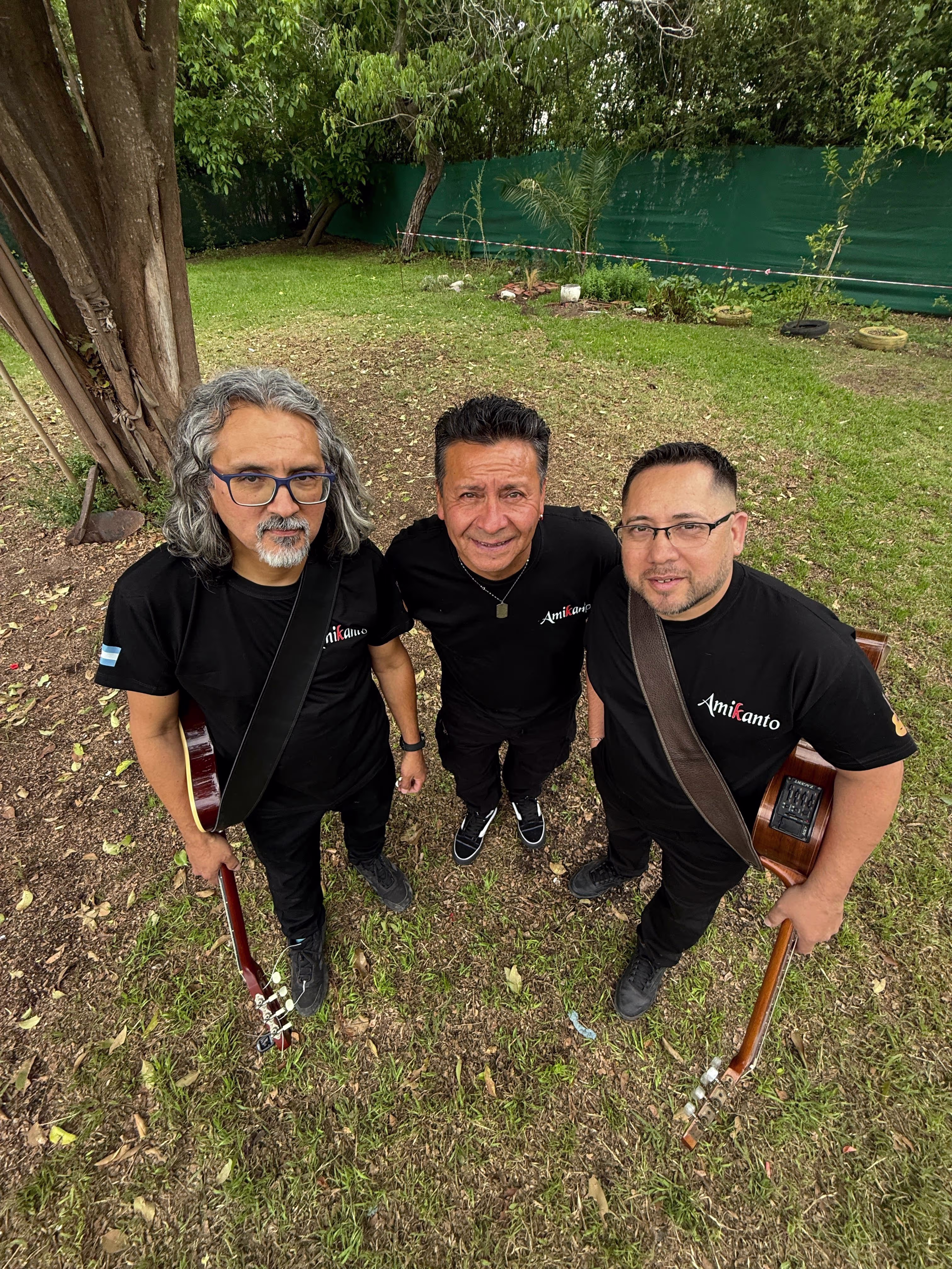 Los tres integrantes de Amikanto posando desde abajo mientras miran hacia la cámara, rodeados de árboles y césped.